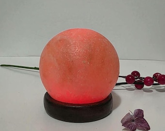 May include: A spherical, glowing orange salt lamp sits on a circular dark brown wooden base. The lamp emits a warm, vibrant light, highlighting the textured, crystalline surface of the salt. A thin stem with small red berries and a purple flower.