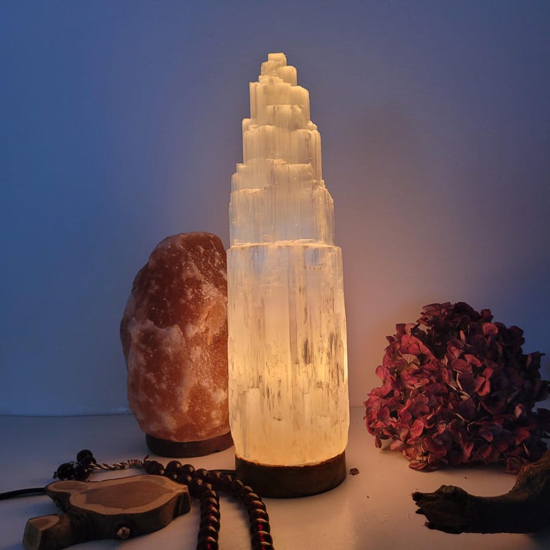 May include: A glowing selenite tower lamp with a layered design, next to a pink Himalayan salt lamp. The selenite lamp emits a soft, warm light. A dark wooden base supports the selenite tower. Dried red flowers and a beaded necklace are also visible.