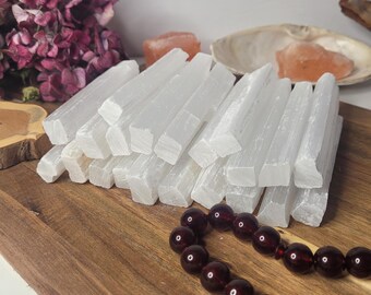May include: A large pile of white selenite sticks, also known as selenite wands, on a wooden surface. The sticks are stacked in a staggered pattern, with the ends of the sticks visible. There is a strand of dark red beads in the foreground.