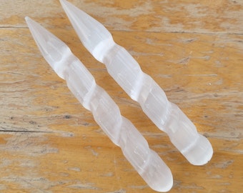 May include: Two white selenite wands with a spiral design. The wands are lying on a wooden surface.