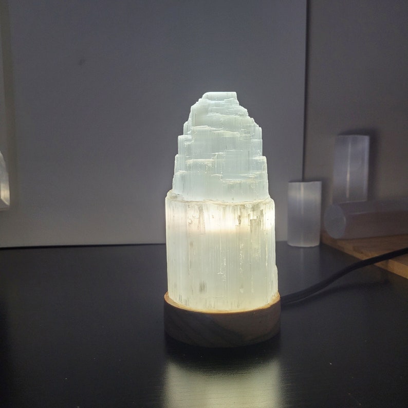 May include: A glowing selenite lamp with a tiered, crystalline structure. The lamp emits a soft, warm light and sits on a round, wooden base. The lamp is approximately 6 inches tall.