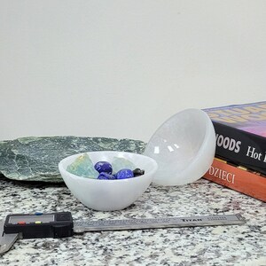 May include: Two white selenite bowls, one filled with blue and green crystals. The bowls are on a granite countertop with a green stone and a metal ruler.
