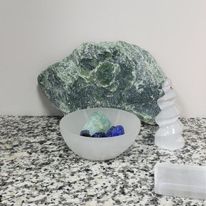 Pu&ograve; includere: Una ciotola bianca opaca piena di cristalli blu e verdi si trova su un piano di lavoro in granito. Uno grande masso verde e una scultura di cristallo bianco sono sullo sfondo.