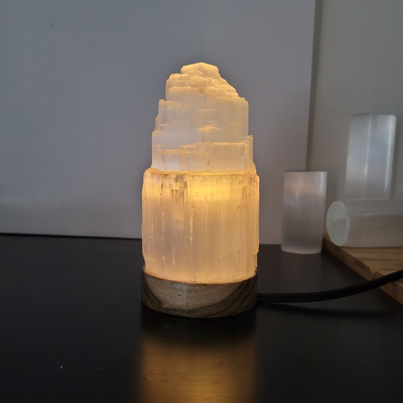 May include: A glowing selenite lamp with a layered, tower-like design. The lamp emits a warm, soft light, and sits on a round wooden base. A black power cord extends from the base. The lamp is a decorative home accent.