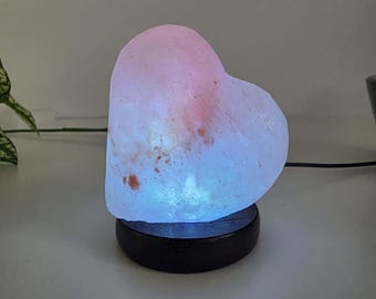May include: A heart-shaped Himalayan salt lamp with a rough, crystalline texture, glowing with a soft pink and blue light. The lamp is mounted on a circular, dark brown wooden base and includes a black power cord extending from the side.