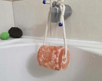 May include: A cylindrical block of pink Himalayan salt with a hole through the center, suspended by a thick white cotton rope with blue plastic tips. The rope is knotted and looped over a silver-toned metal faucet handle in a tiled bathroom.