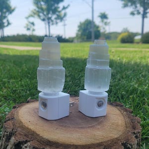 Peut inclure: Deux veilleuses en cristal de sélénite. Chaque lampe a une base blanche et un dessus en cristal de sélénite cylindrique et stratifié. Elles sont présentées sur une souche d'arbre sur fond d'herbe verte et d'arbres.