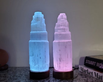 May include: Two selenite crystal lamps, one with blue light and one with pink light, on a dark countertop. The lamps are shaped like a tower with multiple layers. The lamps are sitting on wooden bases.