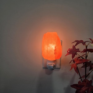 K&ouml;nnte beinhalten: Ein leuchtendes, orangefarbenes Himalaya-Salz-Nachtlicht, das in eine wei&szlig;e Steckdose gesteckt ist. Der Salzkristall hat eine raue Oberfl&auml;che und strahlt ein warmes Licht aus. Im Hintergrund sind r&ouml;tlich-braune Bl&auml;tter zu sehen.