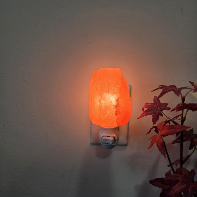 K&ouml;nnte beinhalten: Ein leuchtendes, orangefarbenes Himalaya-Salz-Nachtlicht, das in eine wei&szlig;e Steckdose gesteckt ist. Der Salzkristall hat eine raue Oberfl&auml;che und strahlt ein warmes Licht aus. Im Hintergrund sind r&ouml;tlich-braune Bl&auml;tter zu sehen.