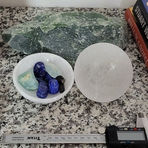 May include: A white selenite bowl filled with blue and green crystals. The bowl is sitting on a gray and white speckled surface. There is a white selenite sphere next to the bowl.
