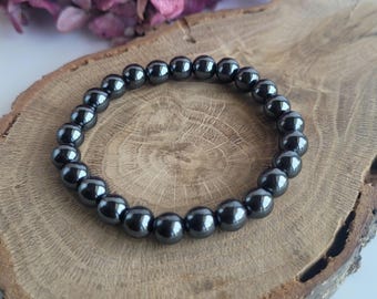 May include: A dark gray beaded bracelet composed of round, polished beads. The bracelet is displayed on a wooden surface with a natural grain pattern. A cluster of purple flowers is visible in the background.