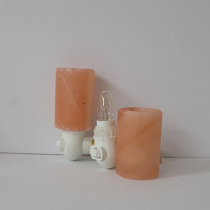 K&ouml;nnte beinhalten: Zwei Himalaya-Salz-Nachtlichter mit einer Gl&uuml;hbirne. Die Salzlampen sind zylindrisch und pfirsichfarben, mit einem wei&szlig;en Kunststoffsockel. Die Gl&uuml;hbirne ist klar und hat eine kleine, abgerundete Form. Die Nachtlichter sind f&uuml;r den Anschluss an eine Steckdose konzipiert.