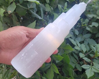 May include: A large white selenite tower with a smooth, striated surface. The tower is held in a hand, with green foliage in the background.
