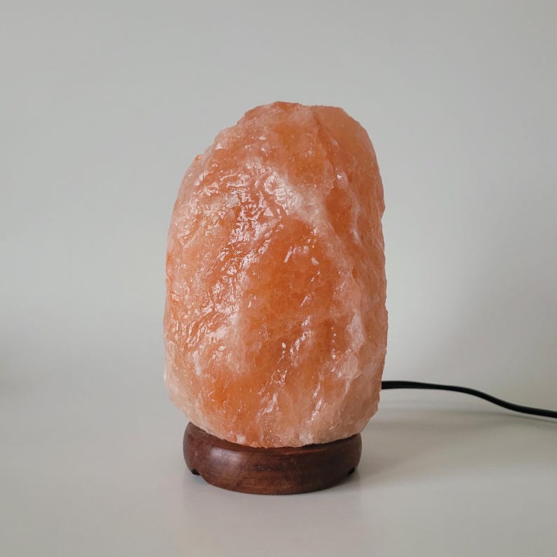 May include: A natural Himalayan salt lamp with a warm orange glow. The lamp is made of a large, irregularly shaped salt crystal with a wooden base.