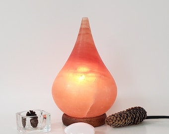 Può includere: Una lampada di sale dell'Himalaya a forma di lacrima con un caldo bagliore arancione. La lampada è su una base di legno e ha un cavo.