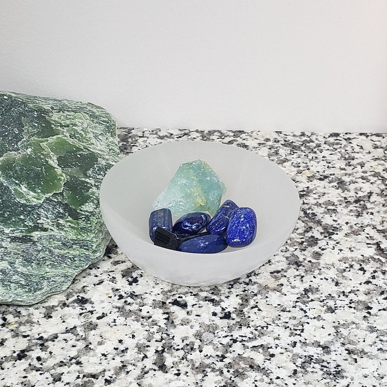 Pu&ograve; includere: Una ciotola bianca piena di cristalli blu e verdi. La ciotola &egrave; su una superficie macchiata di grigio e bianco.