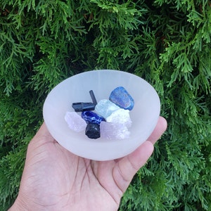 Pu&ograve; includere: Una ciotola di selenite bianca piena di vari cristalli, tra cui cristalli blu, neri e rosa. La ciotola &egrave; appoggiata su una mano.
