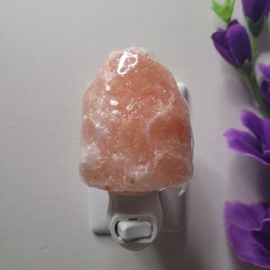 K&ouml;nnte beinhalten: Ein Nachtlicht aus Himalaya-Salz. Die Lampe hat eine hellrosa Farbe und ist wie ein Felsen geformt. Sie ist an eine wei&szlig;e Steckdose angeschlossen. Die Lampe befindet sich neben lila Blumen.