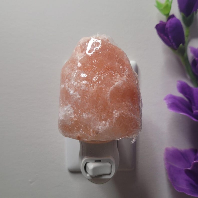 K&ouml;nnte beinhalten: Ein Nachtlicht aus Himalaya-Salz. Die Lampe hat eine hellrosa Farbe und ist wie ein Felsen geformt. Sie ist an eine wei&szlig;e Steckdose angeschlossen. Die Lampe befindet sich neben lila Blumen.