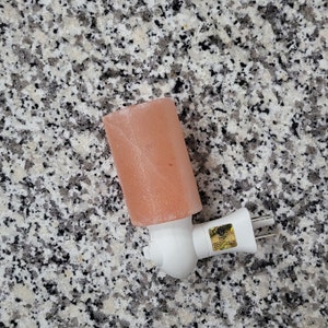 K&ouml;nnte beinhalten: Ein rosa Himalaya-Salz-Nachtlicht mit wei&szlig;em Stecksockel. Der Salzkristall ist rechteckig und hat eine strukturierte Oberfl&auml;che. Der Stecker hat zwei Stifte und einen Sicherheitsaufkleber.