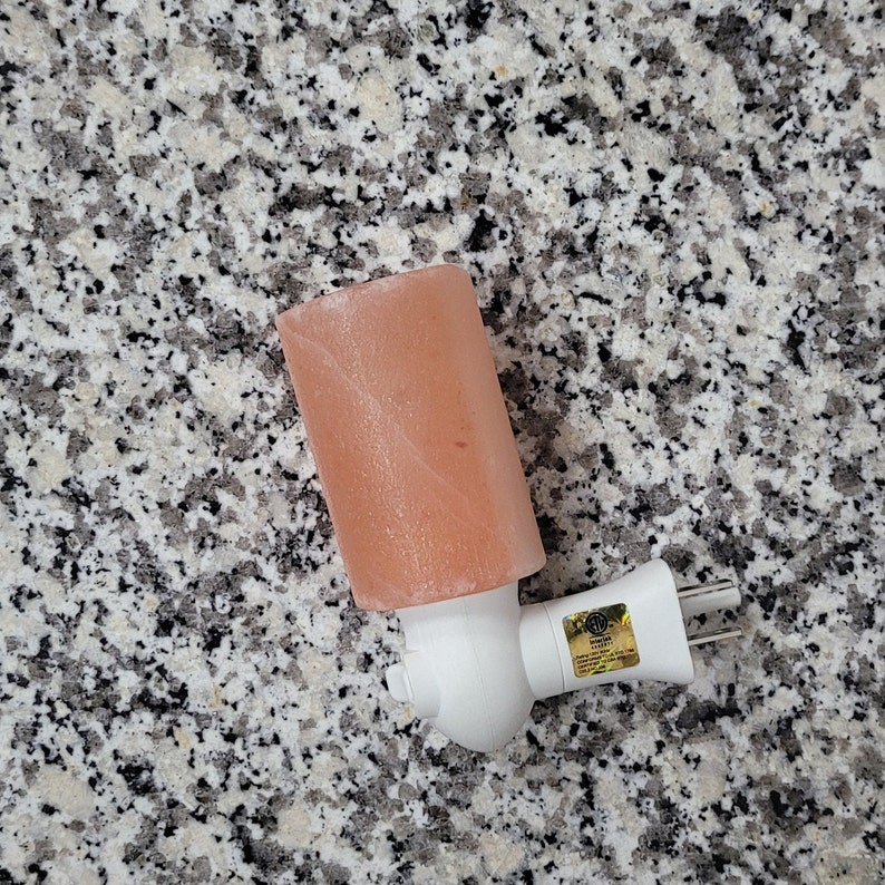 K&ouml;nnte beinhalten: Ein rosa Himalaya-Salz-Nachtlicht mit wei&szlig;em Stecksockel. Der Salzkristall ist rechteckig und hat eine strukturierte Oberfl&auml;che. Der Stecker hat zwei Stifte und einen Sicherheitsaufkleber.