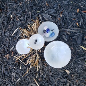 May include: Four white selenite bowls, three small and one large, with a variety of crystals and stones in the center bowl. The bowls are on a bed of black mulch.