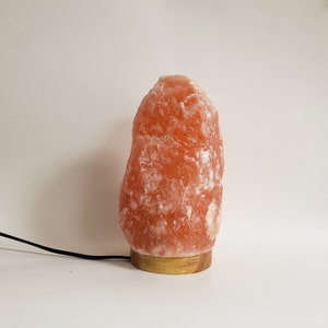 May include: A Himalayan salt lamp with a natural, orange-pink hue. The lamp is on a wooden base and has a cord.