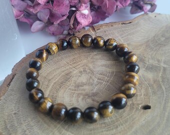 May include: A beaded bracelet made of tiger's eye gemstones. The bracelet features round, polished beads in shades of brown and gold. The bracelet is displayed on a wooden surface, with a backdrop of dried purple flowers.