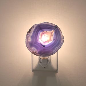 Peut inclure: Veilleuse décorative avec une tranche d'agate violette polie. L'agate présente des bandes de couleur concentriques et une formation cristalline centrale illuminée. La veilleuse se branche sur une prise murale standard et émet une douce lueur chaude.