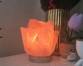 May include: A lotus-shaped Himalayan salt lamp glows with a warm orange light. The lamp sits on a round wooden base. Other decorative items include a gold vase with greenery, a crocheted tulip, and a small potted plant.