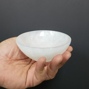 May include: A white selenite bowl, carved from a natural crystal, is held in a hand. The bowl is smooth and polished, with a slightly iridescent sheen.