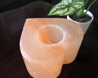 May include: A heart-shaped, light orange Himalayan salt candle holder. The candle holder has a circular opening for a tea light. A small potted plant with green and white leaves is in the background.
