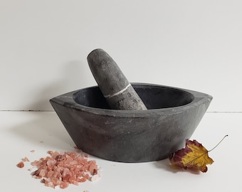 May include: A dark gray stone mortar and pestle set with a smooth, wide bowl and a textured pestle featuring a thin white vein. A small pile of pink Himalayan salt crystals and a single dried yellow and brown leaf sit beside the stone tool.