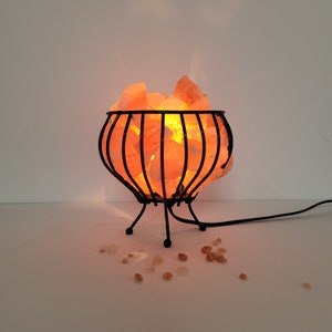 May include: A Himalayan salt lamp with a black metal cage base. The salt crystals are glowing orange from the light inside. The lamp is sitting on a white surface.