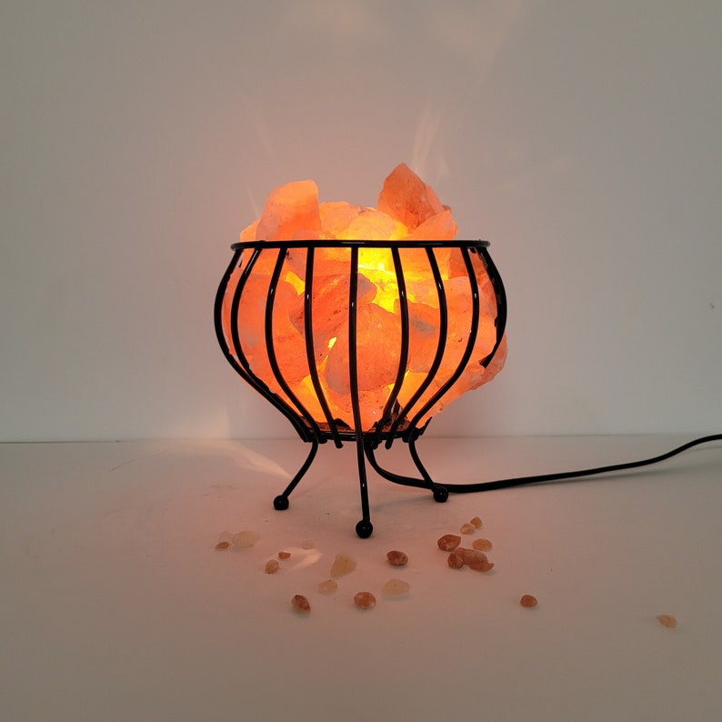 May include: A Himalayan salt lamp with a black metal cage base. The salt crystals are glowing orange from the light inside. The lamp is sitting on a white surface.