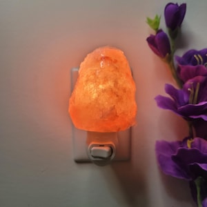K&ouml;nnte beinhalten: Eine beleuchtete Himalaya-Salz-Nachtlampe. Die Lampe ist ein rauer, orangefarbener Kristall, der in eine wei&szlig;e Steckdose gesteckt wird. Im Hintergrund befinden sich lila Blumen.