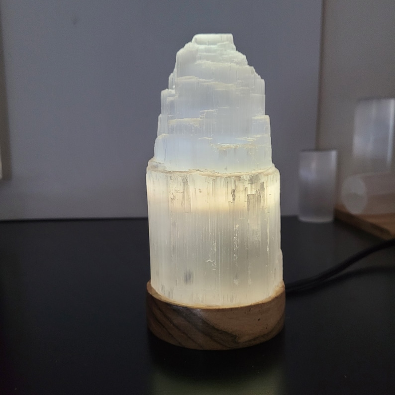May include: A glowing white selenite lamp with a layered, tiered design. The lamp sits on a round, dark wood base. The light emanates from within the selenite, creating a soft, ambient glow. A black power cord extends from the base.