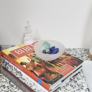 May include: A white selenite bowl filled with blue and green crystals. The bowl is sitting on top of two books, one with the title "Market Math" and the other with the title "Biblia Dla Dzieci".
