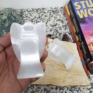 May include: Two white selenite angel figurines. The angel on the left is larger and is being held in a hand. The angel on the right is smaller and is lying on a wooden surface.