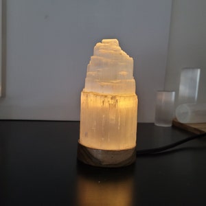 May include: A glowing selenite lamp with a layered, tower-like structure. The lamp emits a warm, soft light, and sits on a wooden base. A black power cord extends from the base. Additional selenite pieces are visible in the background.