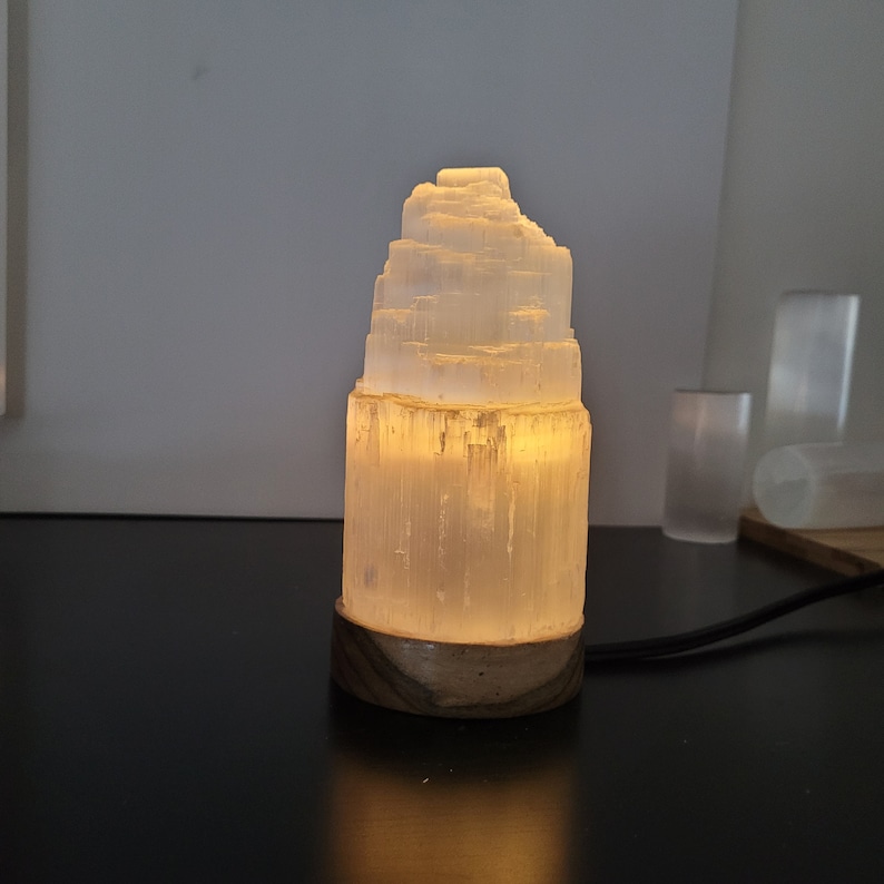 May include: A glowing selenite lamp with a layered, tower-like structure. The lamp emits a warm, soft light, and sits on a wooden base. A black power cord extends from the base. Additional selenite pieces are visible in the background.