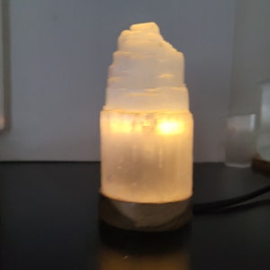 May include: A glowing, white selenite lamp with a layered, tower-like design. The lamp emits a warm, yellow light, creating a soft ambiance. The base is a darker, textured material. A black cord extends from the base.