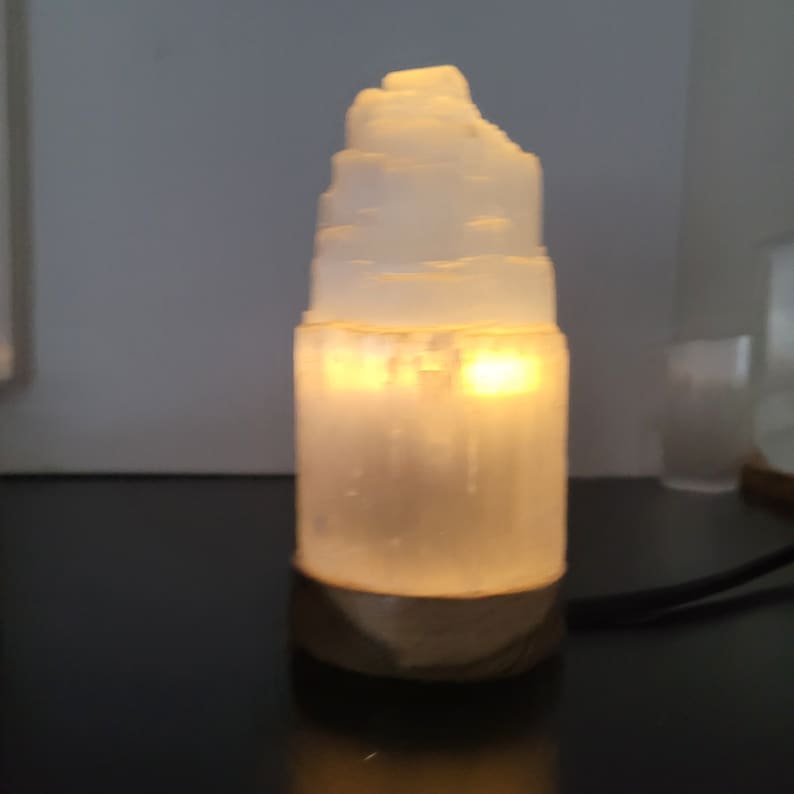 May include: A glowing, white selenite lamp with a layered, tower-like design. The lamp emits a warm, yellow light, creating a soft ambiance. The base is a darker, textured material. A black cord extends from the base.
