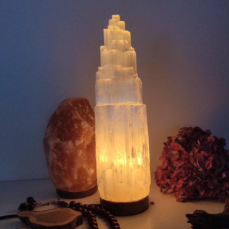 May include: A glowing selenite tower lamp with a layered design, emitting a warm light. Beside it is a pink Himalayan salt lamp and dried red flowers. The lamp is approximately 12 inches tall.