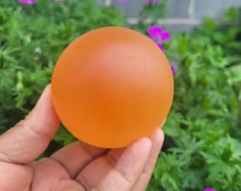 May include: A solid orange sphere, held in a hand, against a backdrop of green foliage and purple flowers. The sphere has a smooth, matte finish and appears to be made of a translucent material. The overall composition is simple and natural.