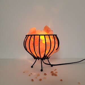 May include: A Himalayan salt lamp with a black metal cage base. The salt crystals are glowing orange from the light inside.