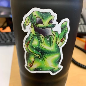 May include: A black tumbler with a sticker of a green cartoon character with a galaxy design. The character has a stitched appearance, with black eyes and mouth. The sticker has a white border.