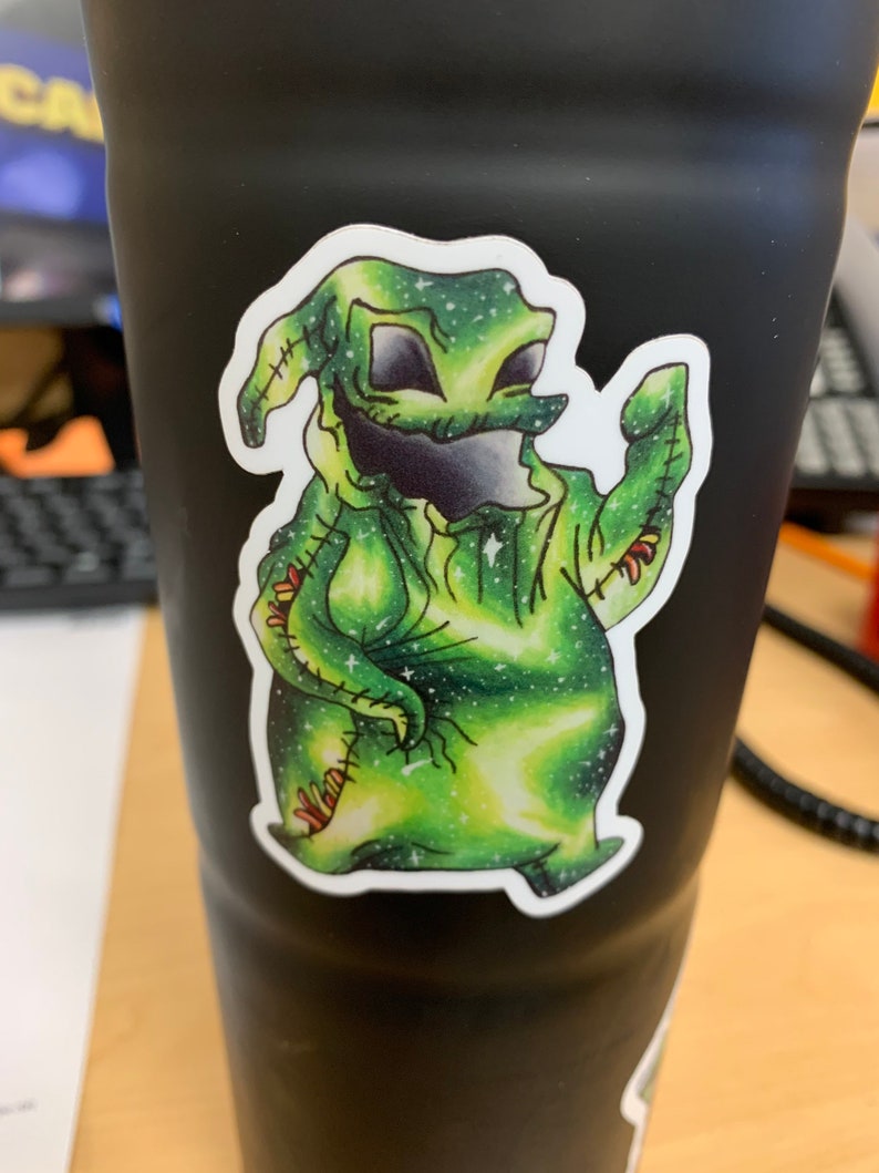May include: A black tumbler with a sticker of a green cartoon character with a galaxy design. The character has a stitched appearance, with black eyes and mouth. The sticker has a white border.