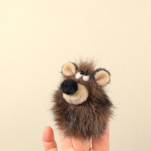 May include: A brown, fuzzy bear face with tan ears and a black nose. The bear has small, black eyes and a light tan muzzle. The bear is held up by a finger.